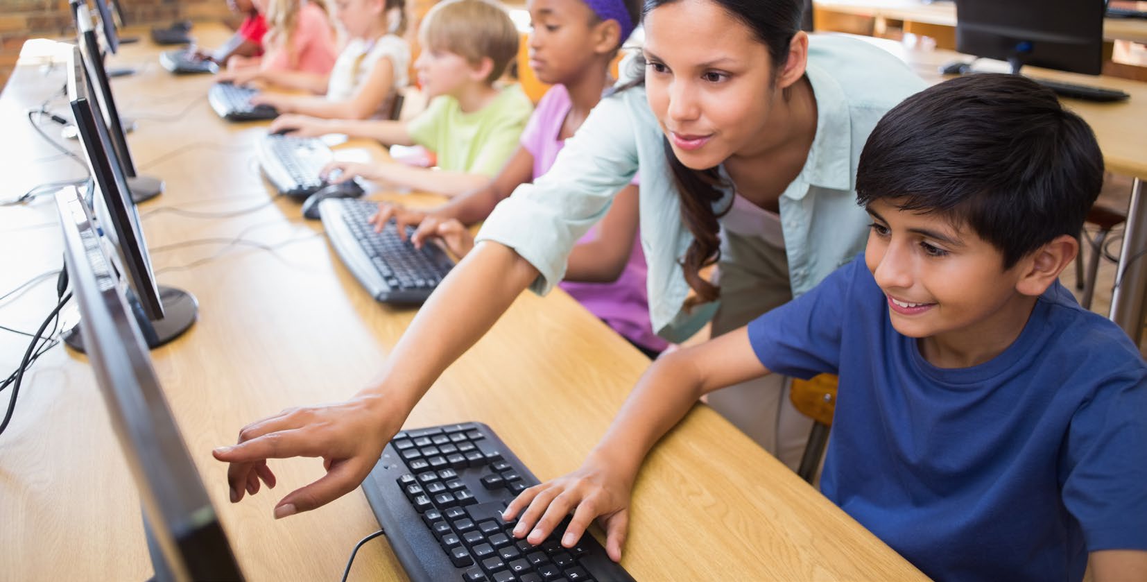 A teacher leaning in to help a young student at a computer in a school computer room