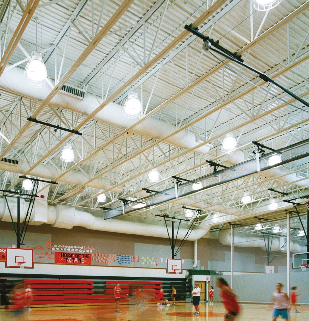 A school sports hall with white steel roof trusses and long fabric ventilation ducts above a basketball court
