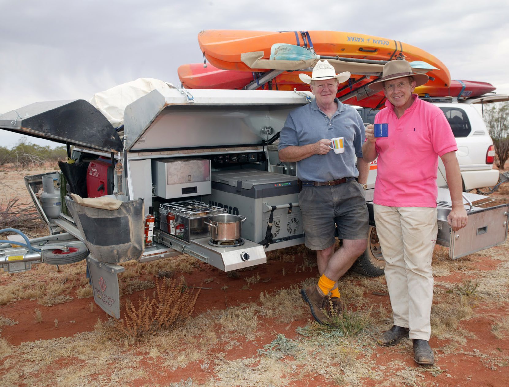 Two men with mugs of tea beside an open camper trailer kitchen in the Australian outback