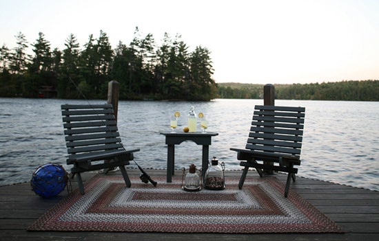 Two wooden chairs and a small table on a lakeside dock, standing on a braided rug